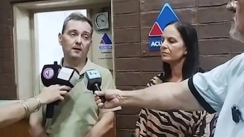 Un hombre y una mujer dan declaraciones a micrófonos de prensa en un edificio de oficinas, con carteles de "ACUSACIÓN" visibles al fondo