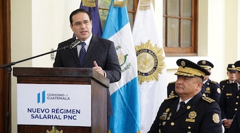 Un hombre con traje y corbata habla en un podio con logo del Gobierno de Guatemala. Detrás hay banderas y un oficial de la Policía Nacional Civil.