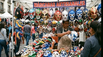 Puesto de mercado lleno de máscaras de lucha libre de múltiples colores, colgando y apiladas. Un hombre con delantal manipula una máscara plateada. Gente en el fondo.