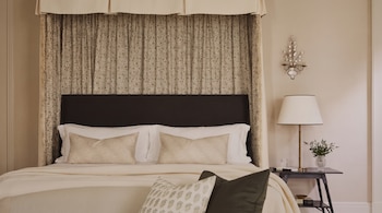 Cama de dosel con cabecero oscuro, sábanas blancas, almohadas decoradas, cortinas estampadas, mesilla de noche con lámpara, jarrón con ramas y aplique de pared.