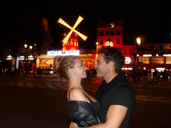 Un hombre y una mujer sonrientes se abrazan de noche en la calle frente a la fachada iluminada del cabaret Moulin Rouge
