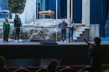 Cuatro personas de pie en un escenario teatral con un fondo de rocas, fachada y ventanas. Una persona sostiene un micrófono en primer plano, de espaldas