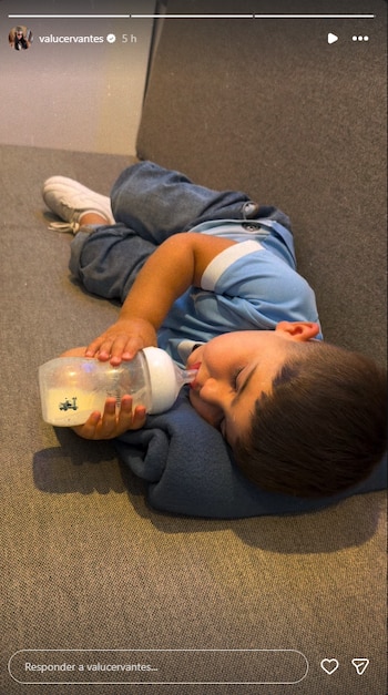 Un niño recostado en un sillón gris bebe de un biberón vistiendo una camiseta celeste de fútbol y pantalones de mezclilla