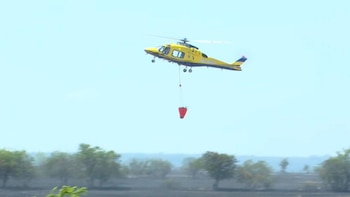 Helicóptero amarillo con balde rojo colgando, volando en un cielo azul claro sobre un terreno con árboles verdes y área oscurecida