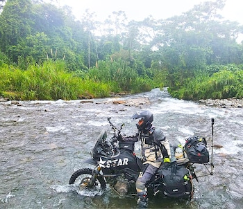 Un hombre con casco y equipo de motocross conduce una motocicleta negra por un río de montaña rocoso, rodeado de densa vegetación verde
