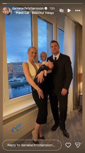 Daniela Christiansson, con un top blanco y falda negra, sostiene a un bebé. Un hombre de traje y un niño pequeño rubio posan junto a una ventana con vista a una ciudad nocturna