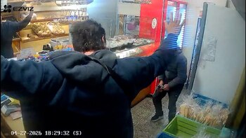 Vista desde atrás de una persona con los brazos levantados dentro de una panadería mientras un asaltante armado la apunta