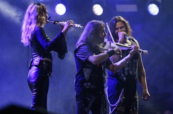 Una flautista, un violinista con un violín blanco y un cantante masculino en un escenario con luces de fondo