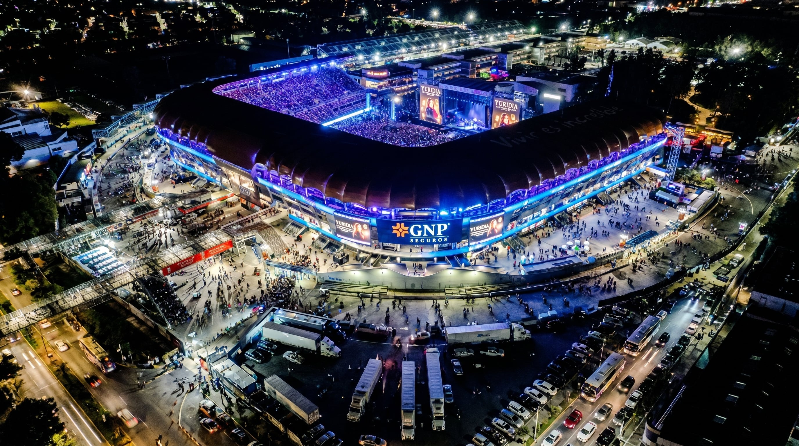 Vista aérea nocturna de un estadio moderno y vibrante, completamente iluminado con luces azules y moradas, que alberga un concierto de Yuridia y muestra el logo de GNP Seguros entre miles de asistentes. (Imagen Ilustrativa Infobae)