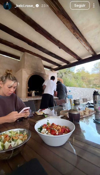 Mulher loira com coque olha para o telefone na mesa de madeira com salada e marido; Ao fundo, dois homens preparam um churrasco de pedra no pátio externo
