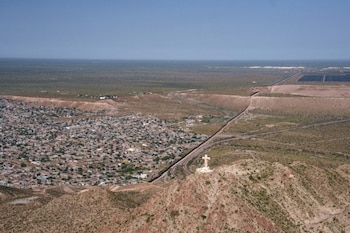 Vista aérea de una ciudad junto a una valla fronteriza en un desierto. Una gran cruz blanca se alza en la cima de una montaña rocosa. Cielo azul