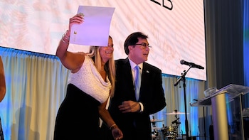 Una mujer sonriente levanta un documento blanco con un logo, junto a un hombre con gafas y traje oscuro que habla en un micrófono en un escenario iluminado