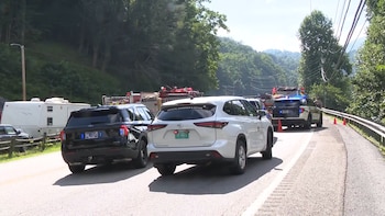 Vehículos de emergencia en una carretera montañosa. Incluye dos coches de policía, un vehículo forense, un camión de bomberos, conos de tráfico y un remolque blanco