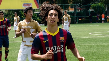 Campo de fútbol de césped verde con varios jóvenes jugadores; uno con camiseta de rayas rojas y azules, otros con camisetas doradas y blancas.