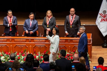 Una mujer en traje blanco saluda con la mano en un escenario. Detrás, una mesa de madera, varias personas con distintivos y una bandera blanca con un logo