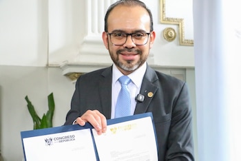 Un hombre con barba y gafas oscuras habla en un podio con micrófonos; detrás, otro hombre de traje azul observa con seriedad