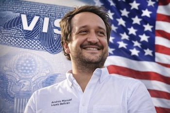 Primer plano de Andrés Manuel López Beltrán sonriente, vistiendo una camisa blanca. Detrás se observan una visa de Estados Unidos y una bandera estadounidense.
