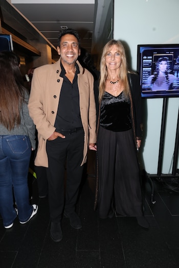 Virginia Elizalde, mujer rubia con blusa de terciopelo negro y pantalón ancho, junto a Coco Fernández, hombre con abrigo camel y camisa negra, sonríen