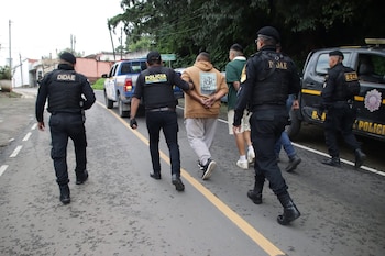 Cuatro agentes policiales caminan por una calle junto a dos hombres esposados que tienen el rostro difuminado