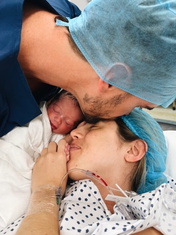 Un hombre con gorro quirúrgico besa la cabeza de un recién nacido. La madre, con gorro y bata, sonríe y sostiene la mano del bebé, en un hospital