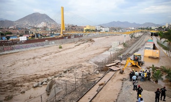 Caudal crecido de un río junto a vías férreas inundadas, una retroexcavadora amarilla, personal de seguridad y un puente atirantado