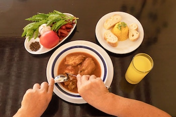Manos sujetan cubiertos sobre un plato de Gulai. Al lado hay arroz amarillo, rebanadas de pan, un vaso de jugo, tomate, ajo, hojas verdes y chiles