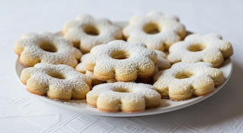 Primer plano de un plato blanco lleno de galletas doradas con forma de flor y un agujero central, cubiertas de azúcar glas, sobre un mantel blanco.