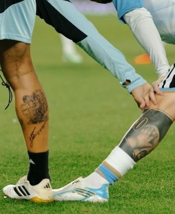 Detalle de las piernas de dos deportistas con tatuajes y calzado deportivo sobre el césped de una cancha de fútbol