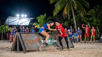 Dos atletas de Exatlón México, uno de azul y otro de rojo, compiten en un obstáculo de madera sobre arena en una playa iluminada de noche. Otros competidores observan.