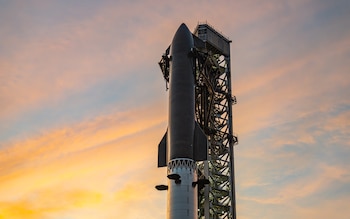 Cohete Starship de SpaceX, gris oscuro y blanco, erguido junto a torre de lanzamiento metálica. Cielo con tonos anaranjados y azules