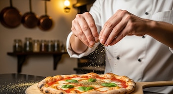 Manos de un chef en uniforme blanco esparciendo orégano seco sobre una pizza redonda con salsa, mozzarella y albahaca fresca en una tabla.