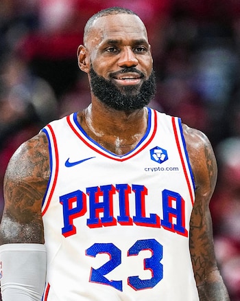 Primer plano de un hombre calvo con barba que viste una camiseta blanca de baloncesto con texto PHILA, número 23, y logos de Nike y Crypto.com