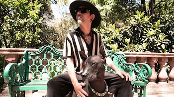 Hombre con sombrero negro y camisa a rayas sentado en un banco verde junto a un perro xoloitzcuintle negro en un parque arbolado.