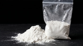 Una bolsa transparente llena de polvo blanco junto a una pila de polvo blanco similar sobre una superficie oscura.