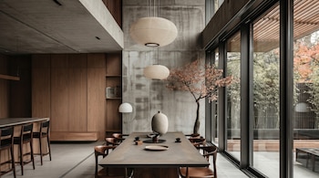 Comedor de estilo moderno con mesa de madera, sillas y dos lámparas colgantes redondas. Un árbol interior y ventanales con vista a un jardín de bambú.