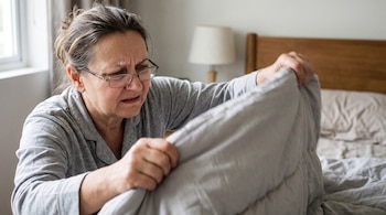 Una mujer con gafas y pijama gris sostiene un edredón en un dormitorio junto a una cama de madera.