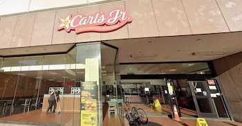 Una vista exterior de un restaurante Carl's Jr con su cartel rojo y amarillo, ventanas de cristal, un empleado barriendo y bicicletas estacionadas afuera