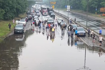 Vista aérea de una autopista con un carril cubierto de agua, decenas de vehículos detenidos y personas de pie en el asfalto y los márgenes arbolados