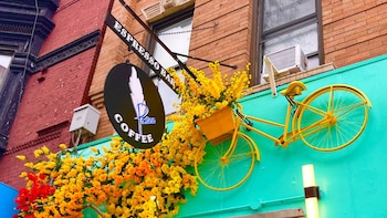 Fachada de edificio de ladrillo y turquesa. Letrero negro "Espresso Bar Coffee Poetica". Bicicleta amarilla, canasta con flores amarillas y naranjas