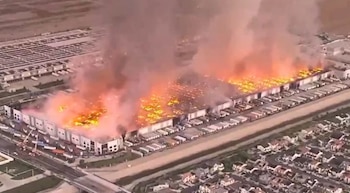 Vista aérea de un gran almacén industrial blanco envuelto en intensas llamas y denso humo gris y naranja, con camiones estacionados y casas cercanas