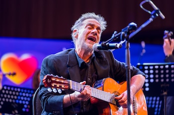 Un hombre canta en micrófono y toca guitarra acústica en un escenario iluminado de azul con una figura de corazón