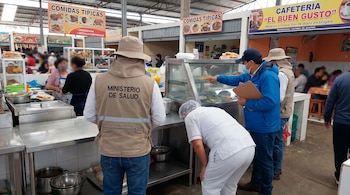 Personeros del Ministerio de Salud inspeccionan un puesto de comida en un mercado. Un oficial señala alimentos en una vitrina; otro lleva chaleco distintivo.