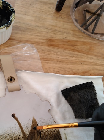 Mano que usa un pincel para pintar una pieza de cuero junto a un recipiente con tinta y una esponja sobre una mesa de madera