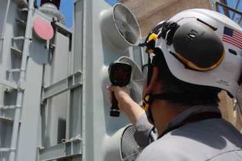 Hombre con casco blanco que lleva bandera de Estados Unidos. Sostiene una cámara térmica apuntando a un transformador eléctrico con varios ventiladores