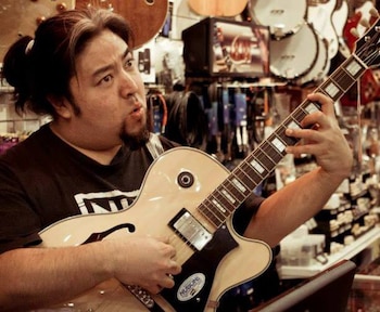 Hombre con barba de chivo y cabello recogido toca una guitarra eléctrica color crema con incrustaciones rectangulares en el mástil dentro de una tienda de música