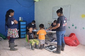 En una sala, adultos y niños participan en un chequeo médico. Se observa a una niña sentada, personal médico en una mesa y una báscula en el suelo.