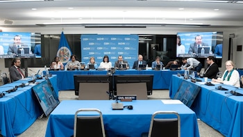 Sala de conferencias con mesas azules, personas sentadas, pantallas de video, una bandera, una pancarta, micrófonos, ordenadores y botellas de agua
