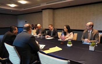 Personas con traje formal conversan alrededor de una mesa de reuniones donde hay banderas pequeñas de Guatemala y Estados Unidos