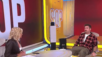 Migue Granados, con camisa a cuadros y barba, sentado en un sillón gris frente a una mujer rubia en un set de televisión con luces de neón amarillas