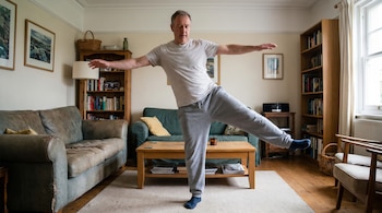 Hombre de mediana edad en camiseta gris y pantalones deportivos grises, de pie sobre una pierna con los brazos extendidos en un salón con sofás, mesa y estanterías.